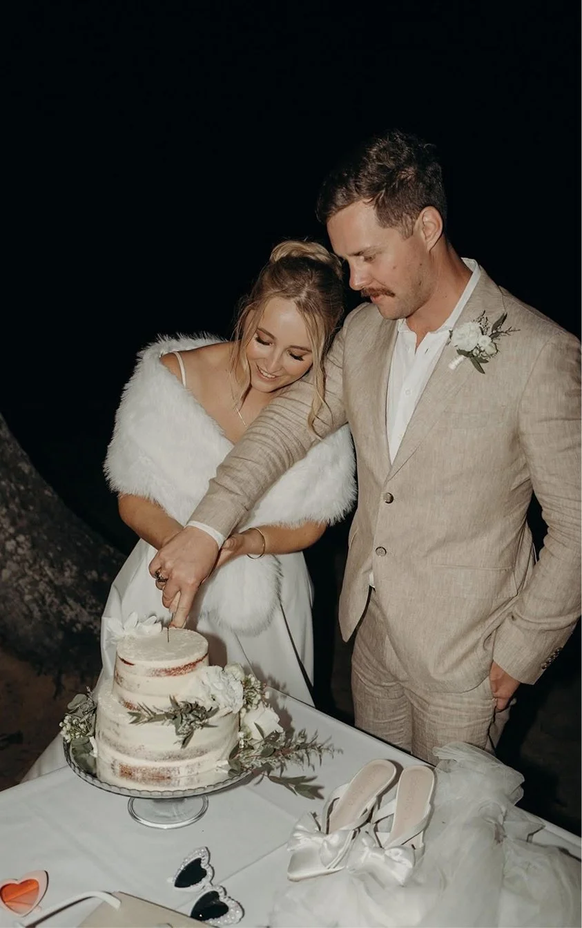 A couple in wedding attire cutting a wedding cake outdoors at night.