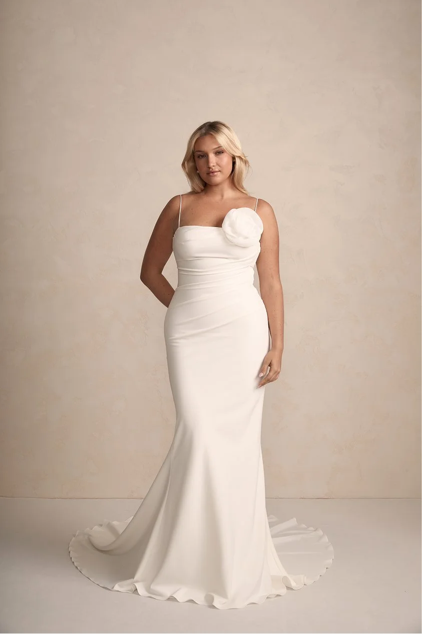 A woman wearing a white, strapless, floor-length wedding dress with a large fabric flower detail on the upper left side, standing against a plain, beige background.