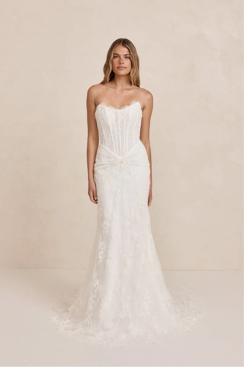 Young woman in a strapless white wedding dress with lace details, standing against a neutral background.