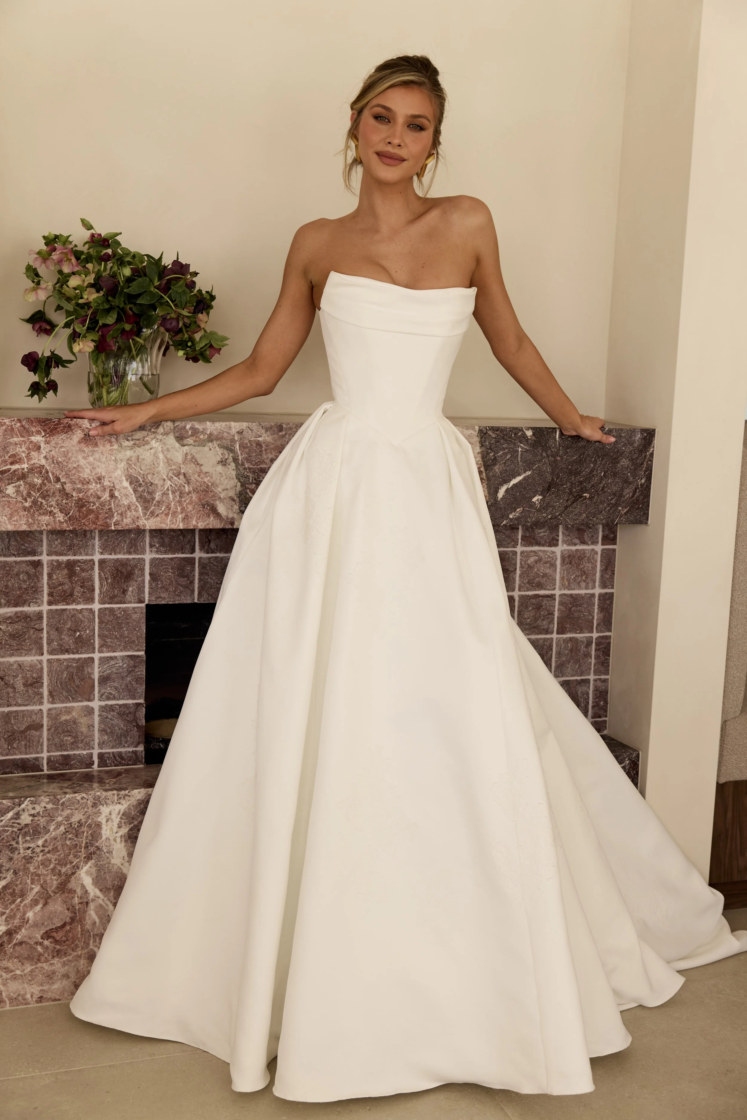 Model wearing the Monroe wedding gown by MWL, featuring a strapless sculpted bodice with a softly draped neckline, basque waist and full A-line skirt with subtle lace detailing, photographed beside a statement marble fireplace.