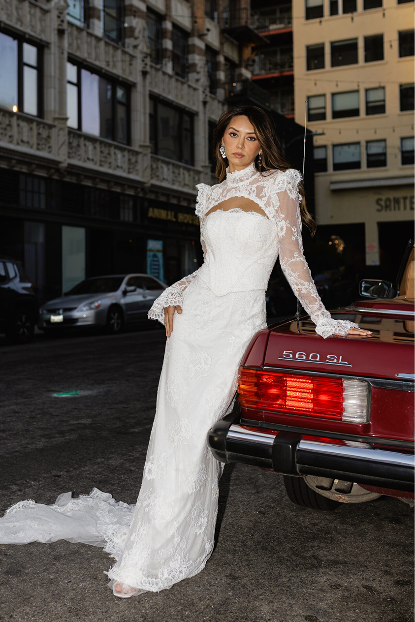 A woman dressed in a white lace wedding gown standing next to a red vintage Mercedes-Benz 560 SL in an urban street setting during twilight.