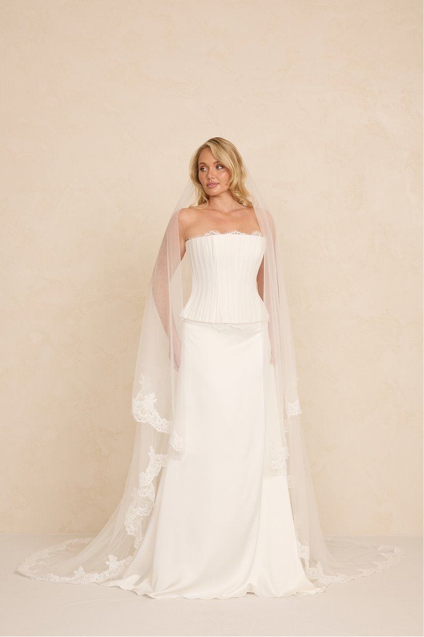 A woman in a wedding dress with a veil, standing against a neutral background.