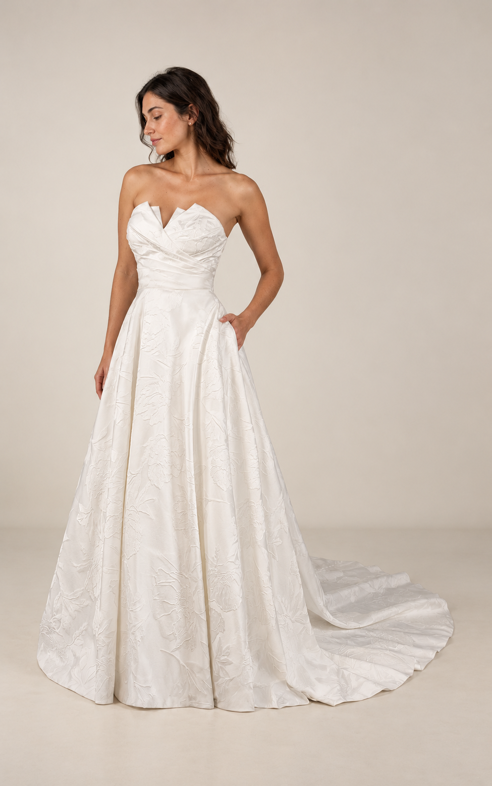 A woman wearing a strapless white wedding dress with a textured floral pattern, standing against a plain background, with her hand in her pocket and her eyes closed.