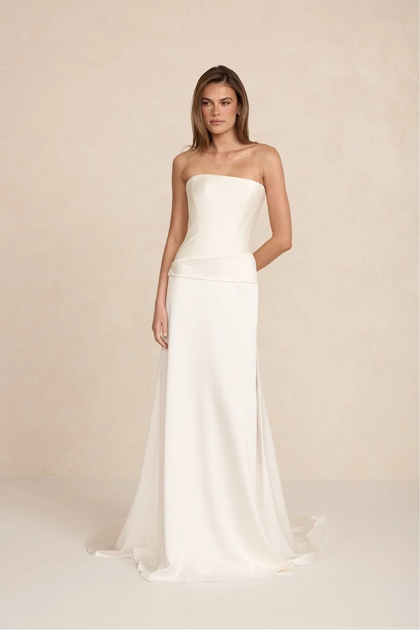 A woman wearing a strapless white wedding dress with a fitted bodice and a long flowing skirt, standing against a plain beige background.