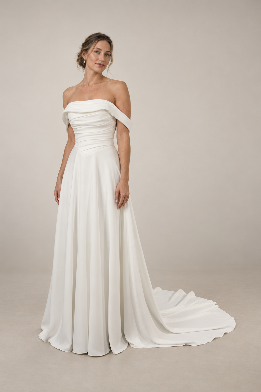 A woman wearing a strapless white wedding gown with off-the-shoulder sleeves, standing in front of a plain white wall.