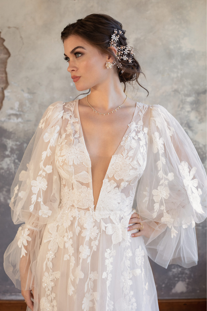 Woman in a wedding dress with floral lace details and puffed sheer sleeves, wearing floral hair accessories and jewelry, standing against a textured wall.