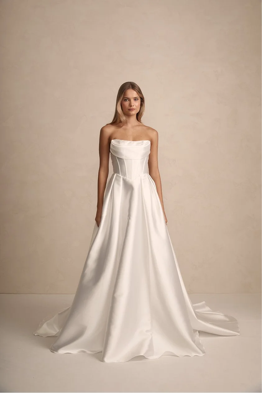 A young woman in a strapless white satin wedding dress standing against a plain beige background.