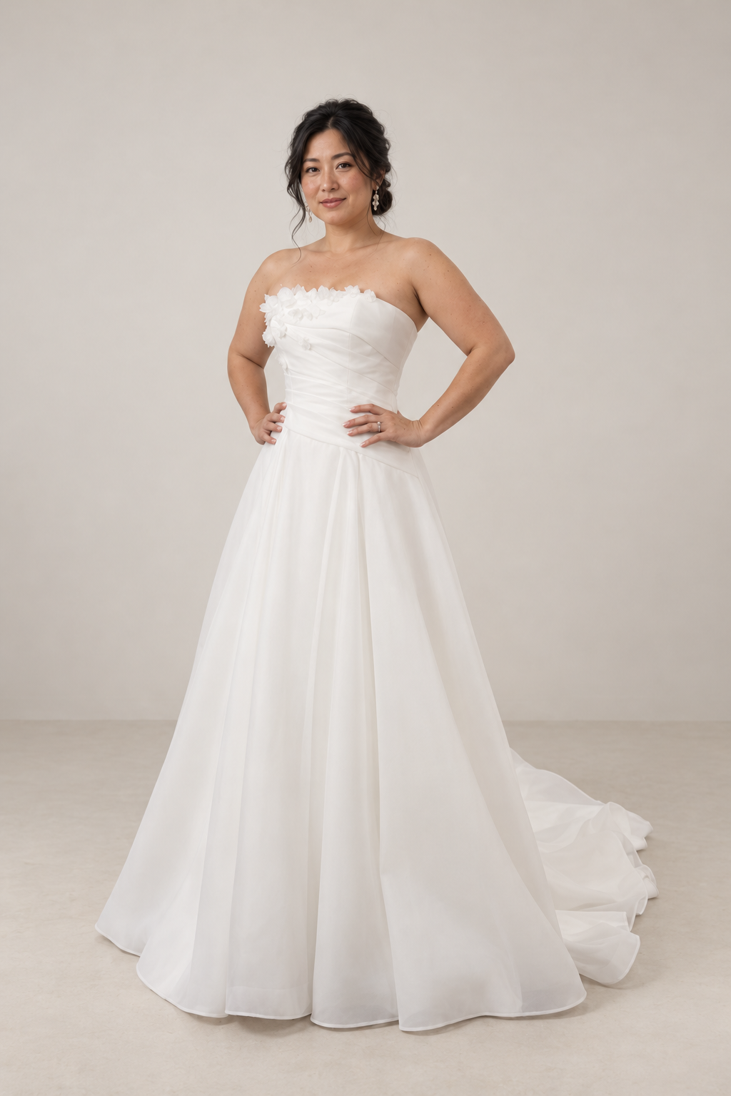 A woman in a white wedding gown with floral details on the strapless bodice, standing against a plain grey background.