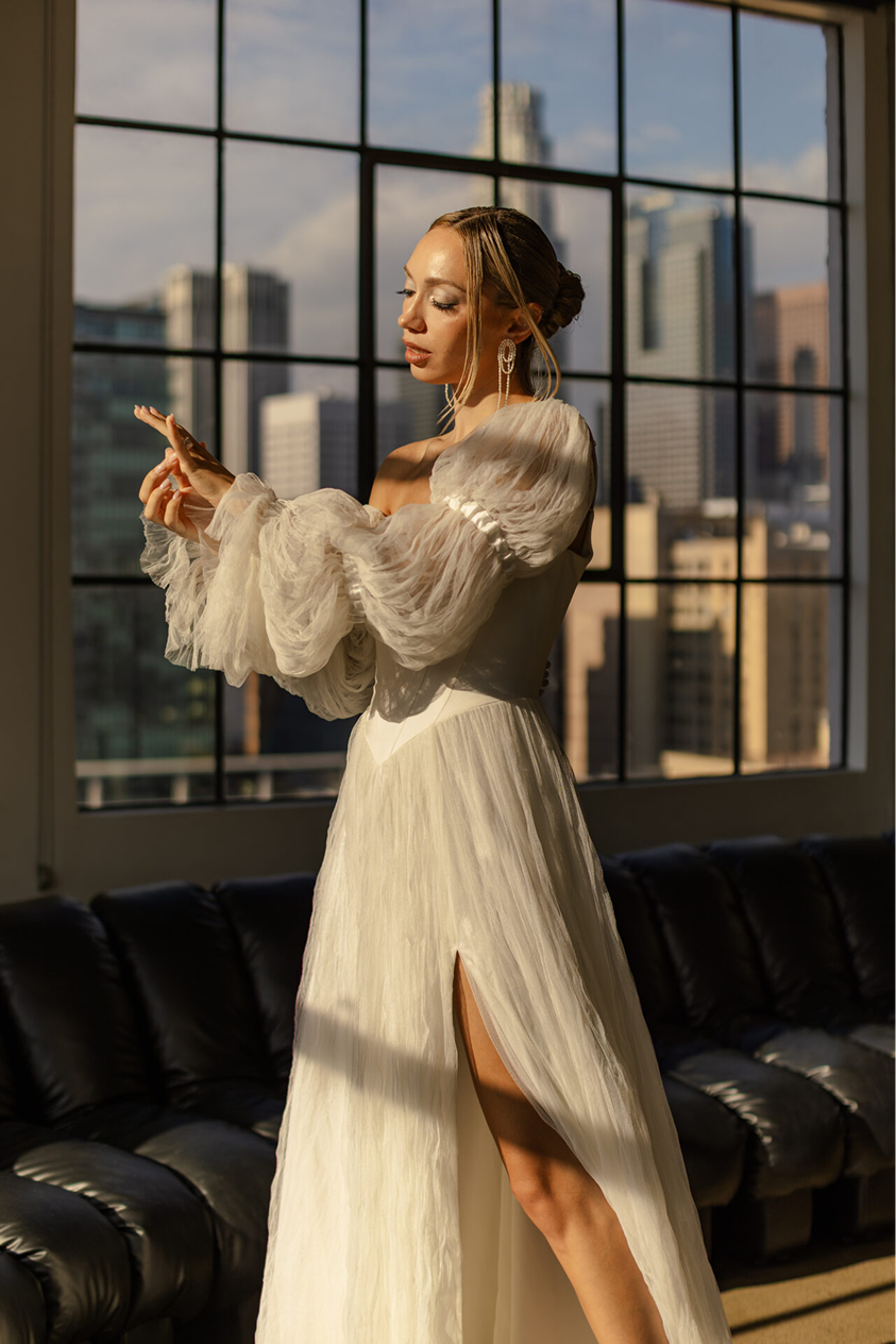 A woman in an elegant off-shoulder cream dress with puffy sleeves and high slit, standing inside near a large window with a city skyline in the background, adjusting her jewelry.