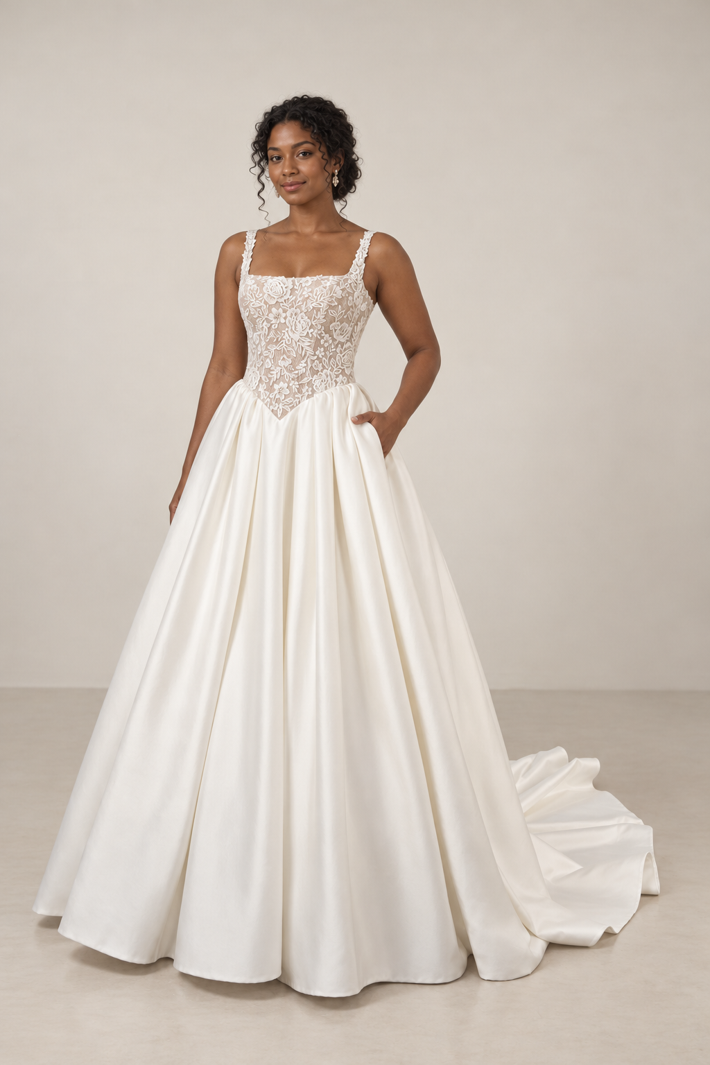 A woman in a white wedding gown with a lace bodice and a full satin skirt, standing with one hand in her pocket against a plain gray background.