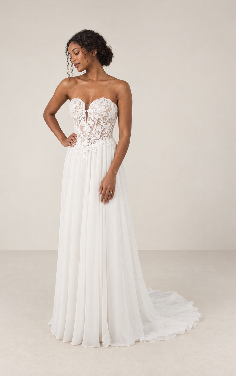 A woman wearing a white strapless gown with a lace bodice and a flowing pleated skirt, standing indoors against a plain background.