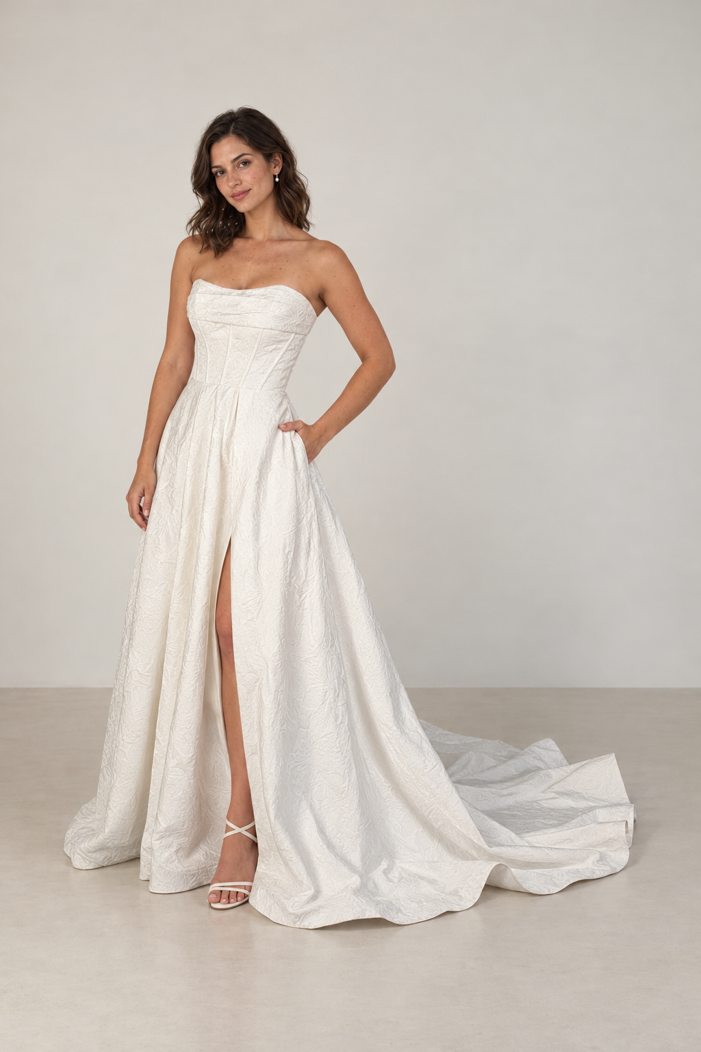 A woman in a strapless white wedding gown with a high slit, standing with one hand in her pocket, against a plain gray background.