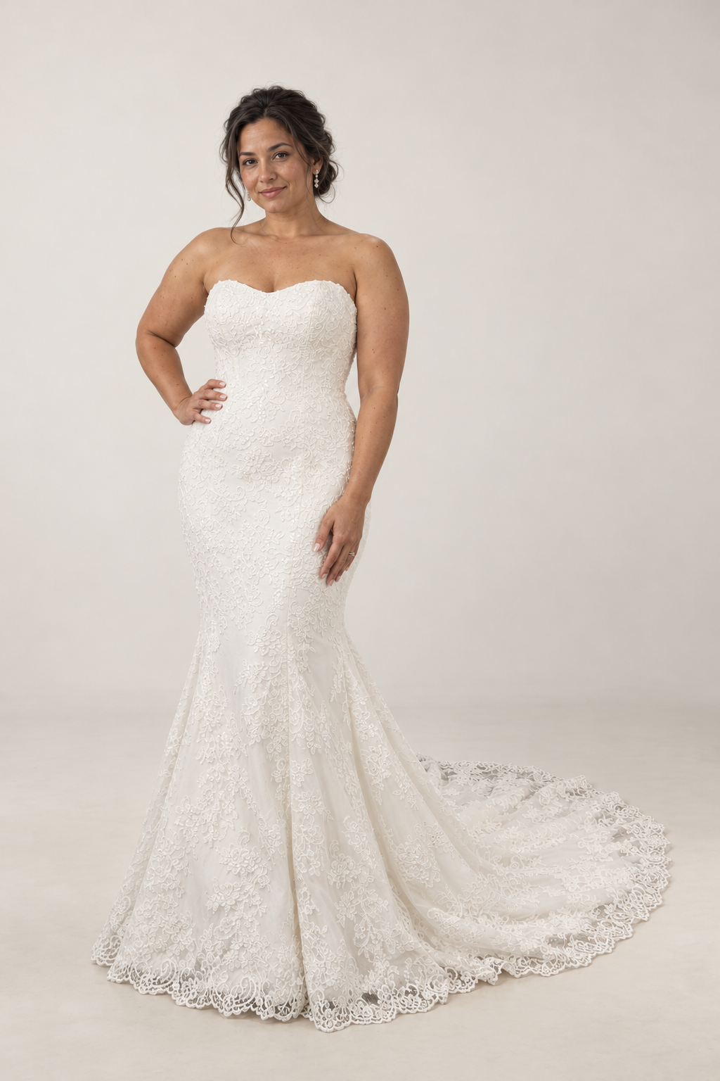 A woman in a strapless white lace wedding gown with a train, standing indoors on a gray floor against a plain beige wall.