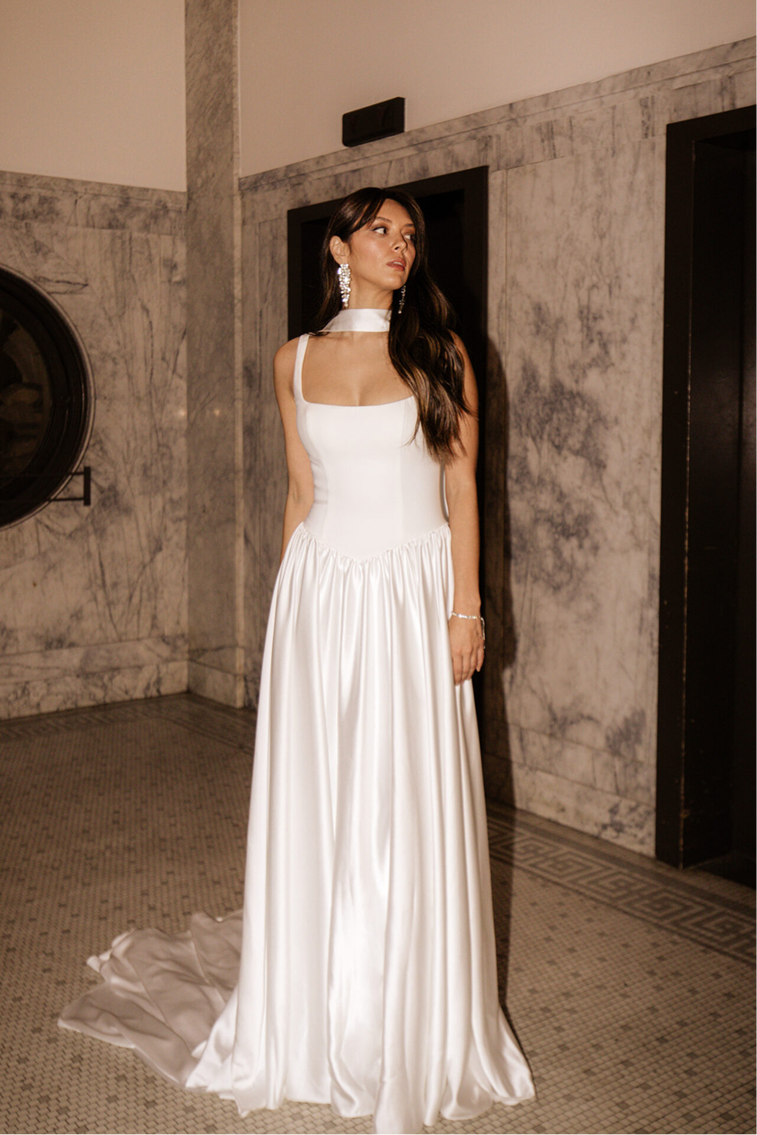 A woman in a long white dress with a fitted bodice and a gathered skirt standing in front of a marble wall.