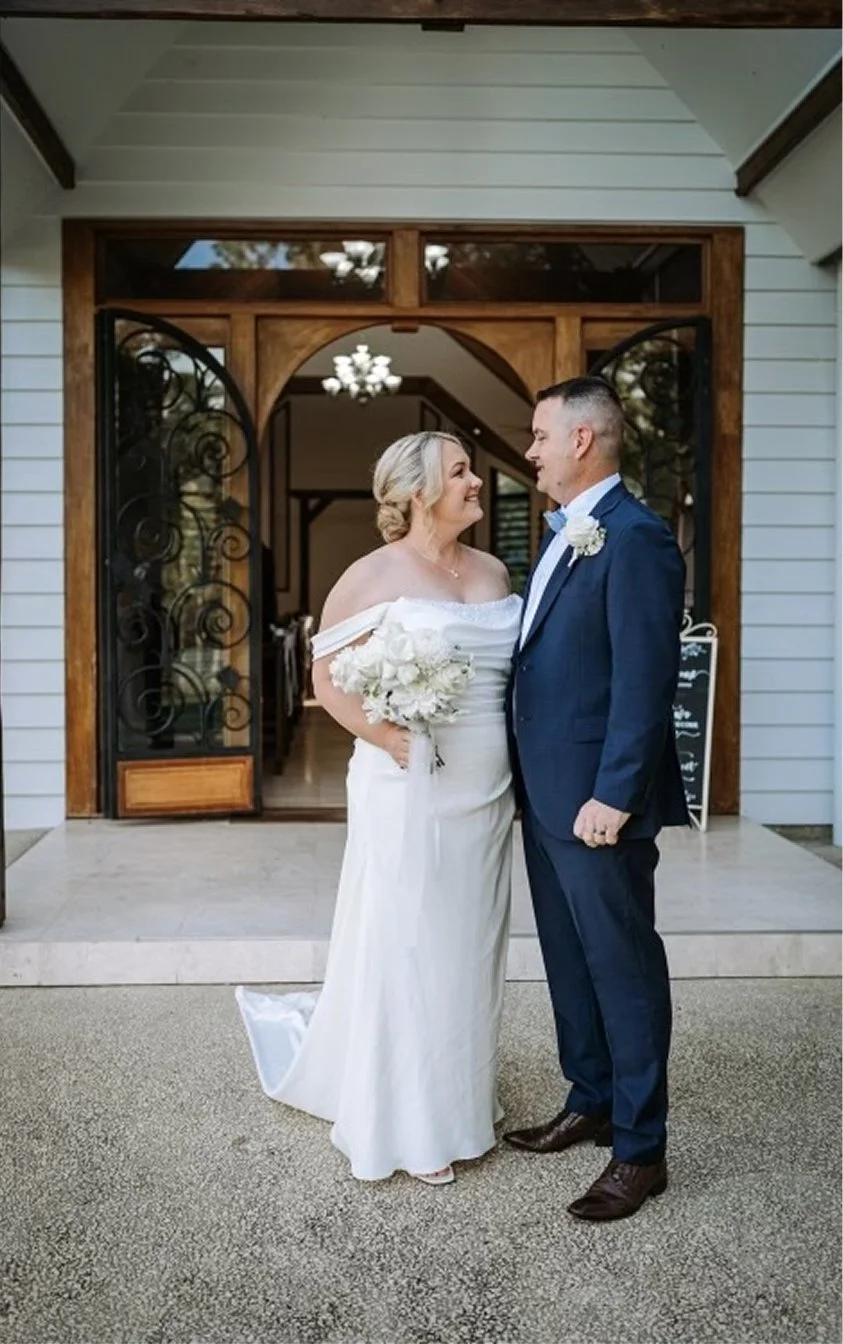 Bride and groom standing face to face outside a building with wooden and iron gate, smiling at each other, with the bride in a white wedding dress holding a bouquet of white flowers and the groom in a dark blue suit with a white boutonniere.