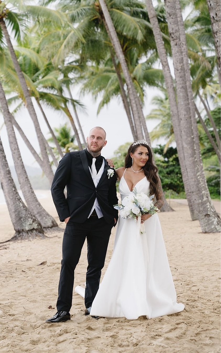 A bride and groom standing on a sandy beach with tall palm trees in the background, bride holding a bouquet of white flowers, bride wearing a white wedding gown and tiara, groom in a black tuxedo with a white shirt, in a tropical outdoor setting.