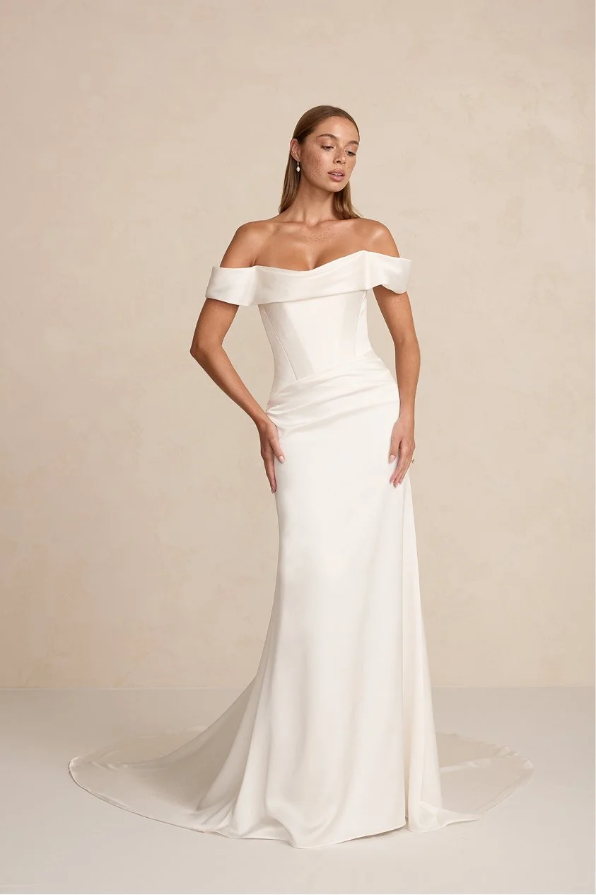 A woman wearing an off-shoulder white wedding gown with a fitted bodice and train, standing against a beige wall.