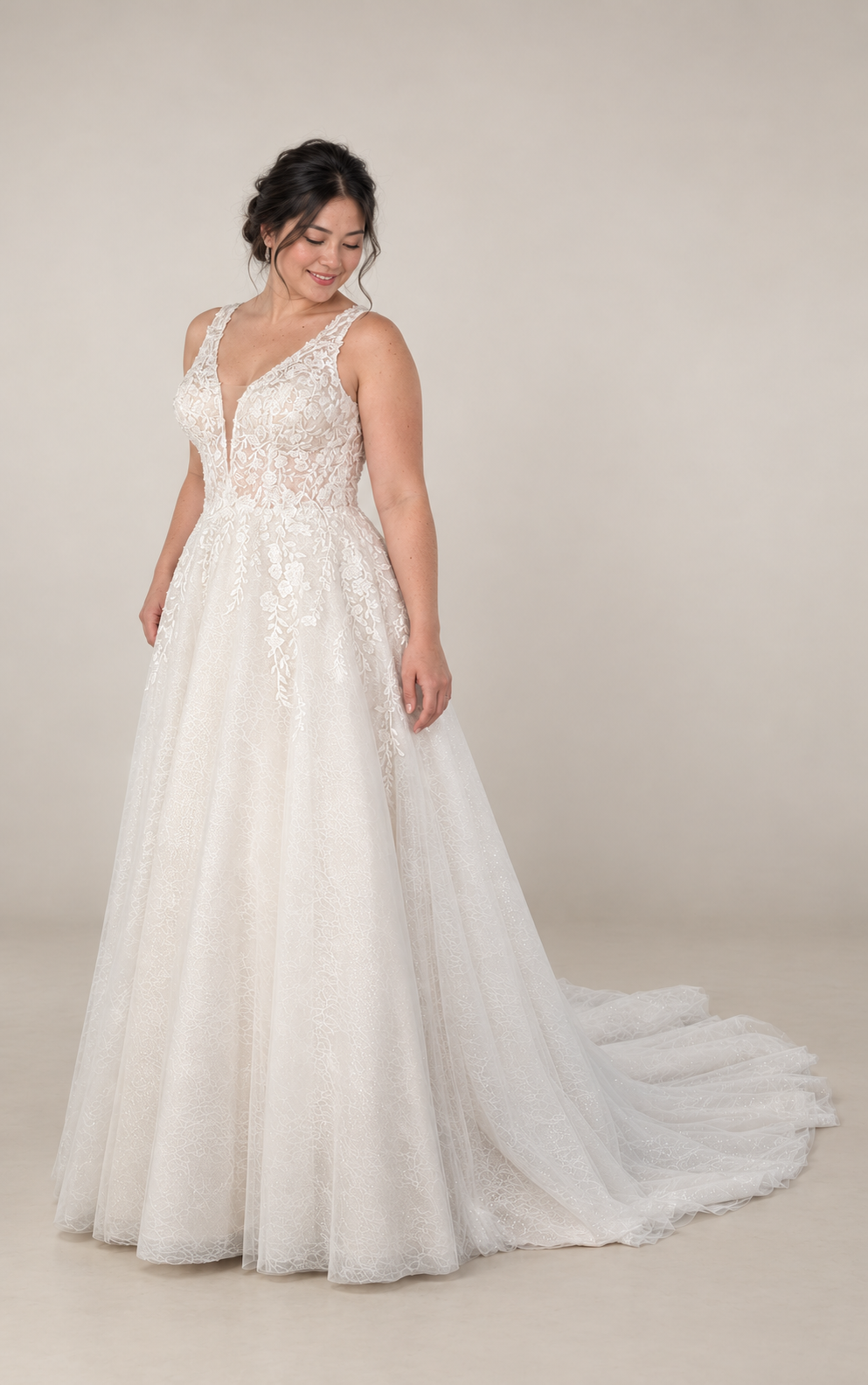 Bride in a white wedding dress with lace details, smiling and looking down, standing on a gray floor against a plain background.