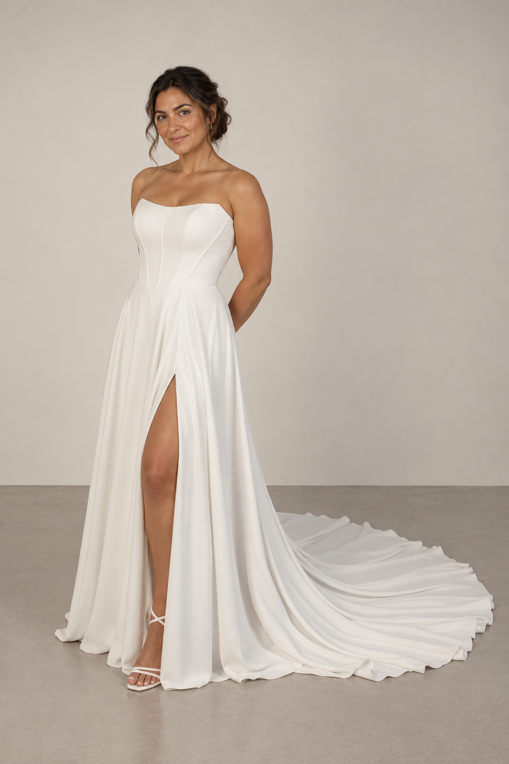 A smiling woman in a strapless white wedding dress with a high slit, standing on a plain gray floor against a neutral background.