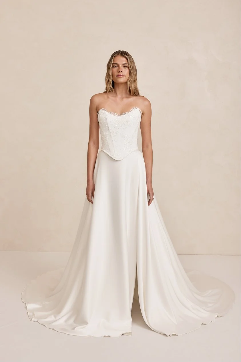 A woman in a strapless white wedding gown with a lace bodice and long satin skirt standing against a neutral background.
