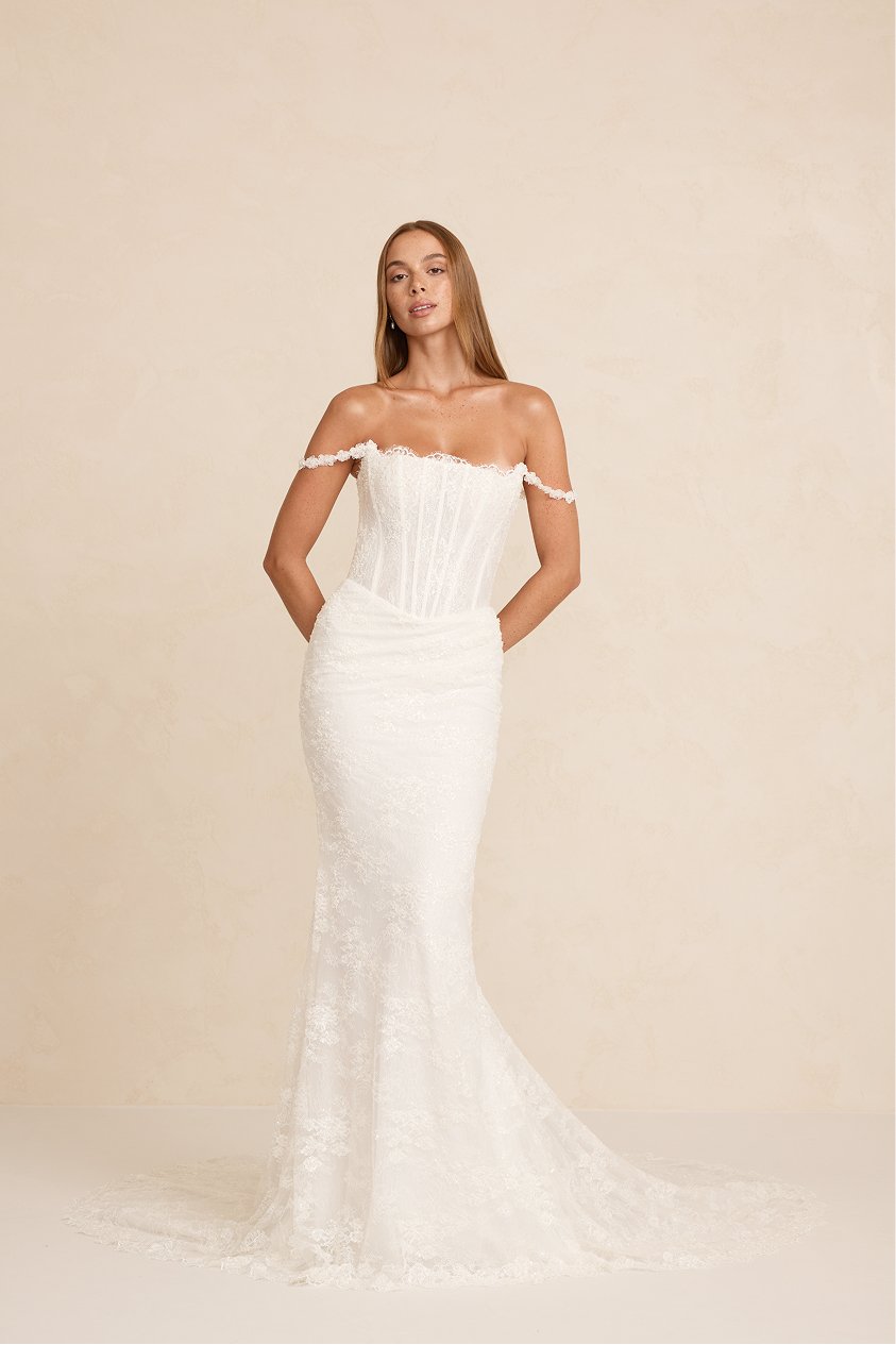 A woman wearing a white off-the-shoulder wedding dress with lace details, standing against a neutral background.