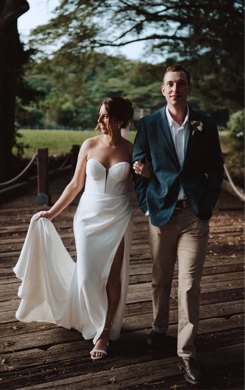 Bride and groom walking on a wooden bridge outdoors during their wedding day. The bride is wearing a white strapless wedding dress with a slit, and the groom is in a navy blazer, white shirt, and beige pants.