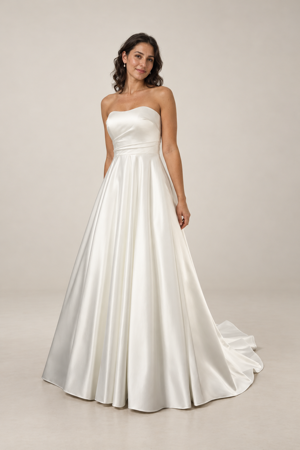 A woman in a strapless white satin wedding dress with a fitted bodice and full skirt, standing against a plain wall.