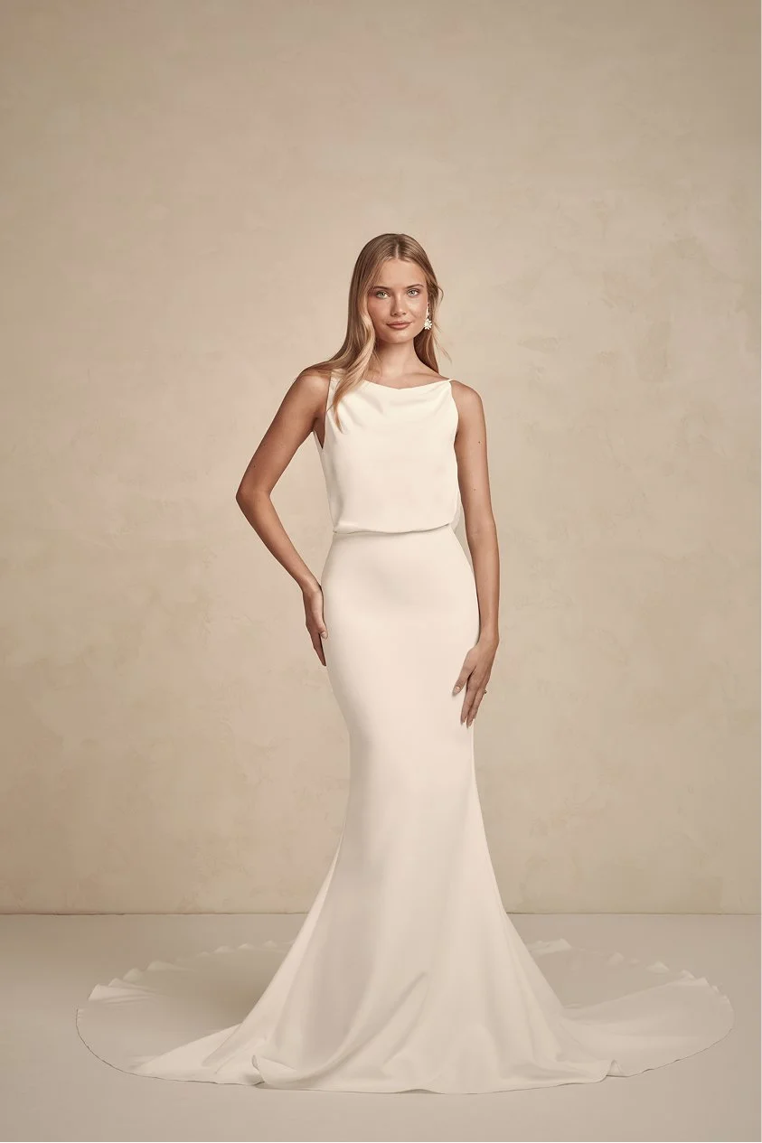 A woman in a sleeveless white wedding gown with a fitted silhouette, standing against a plain beige background.