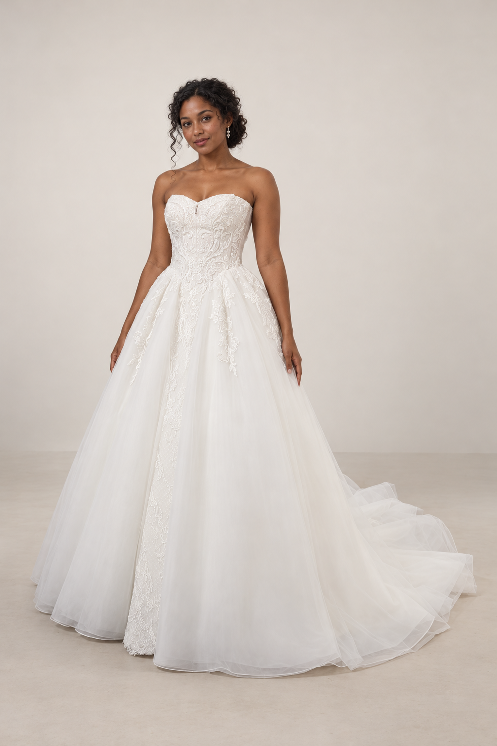 A woman wearing a white strapless wedding gown with lace details, standing indoors against a plain wall.