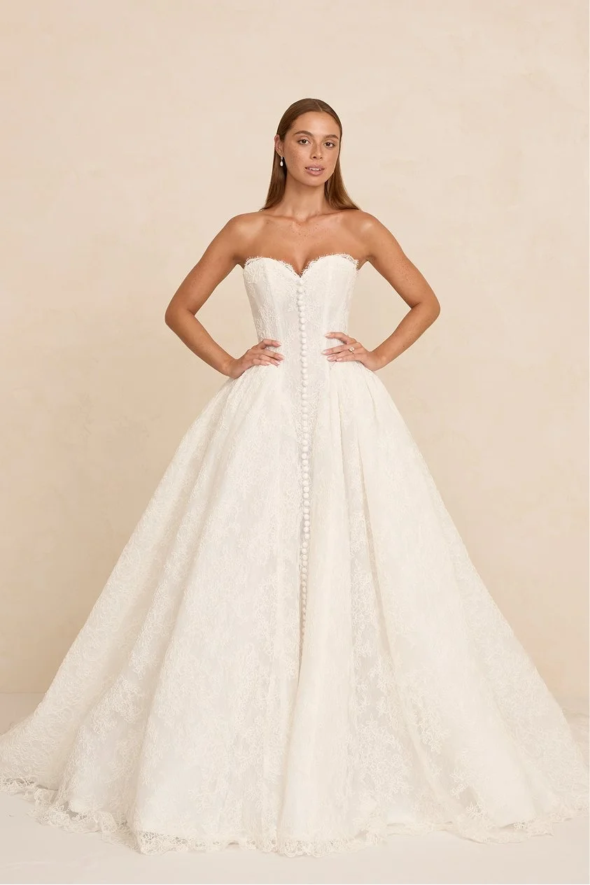 A woman wearing a strapless white wedding gown with lace details and buttons down the front, standing with her hands on her hips against a neutral background.