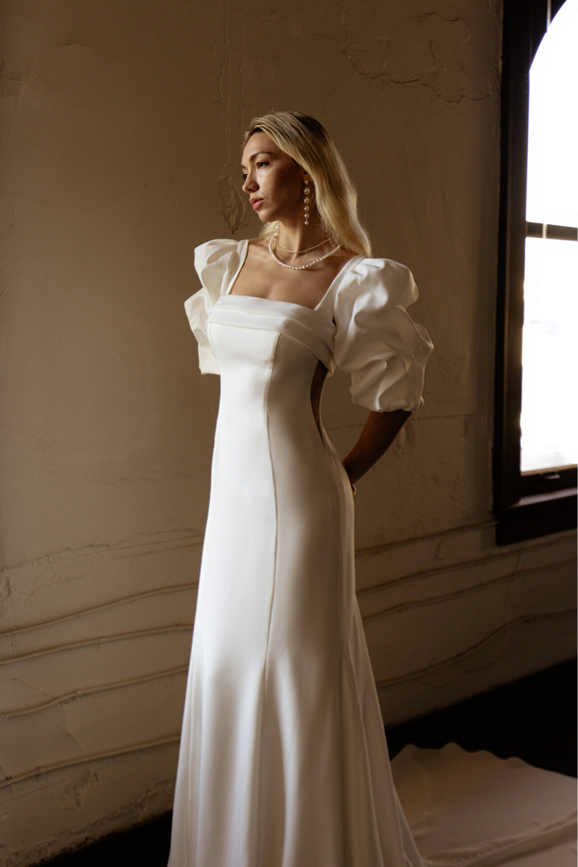 A woman in a white wedding dress with puffed sleeves and pearl jewelry stands by a window in a dimly lit room.