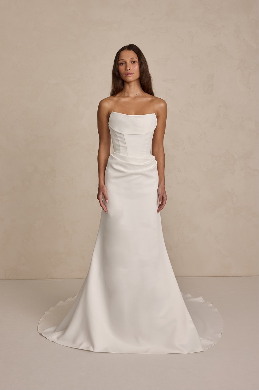 A woman in a strapless white wedding dress standing against a neutral background.