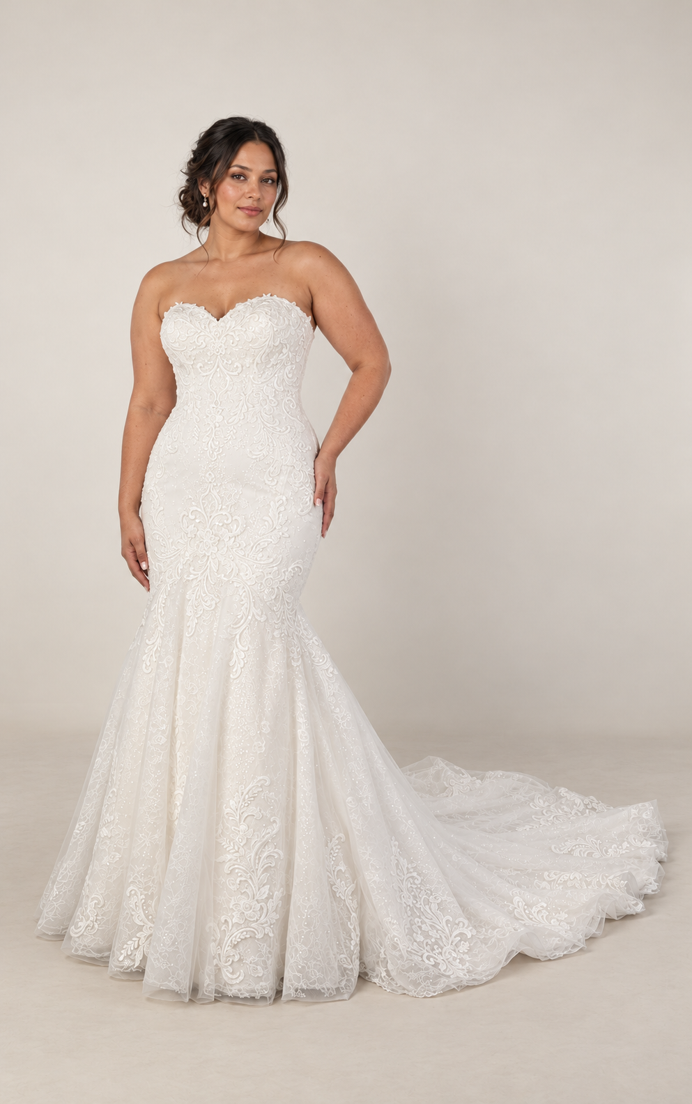 A woman in a strapless, white wedding dress with lace embroidery, fitted at the waist, flaring into a long train, standing against a plain gray background.