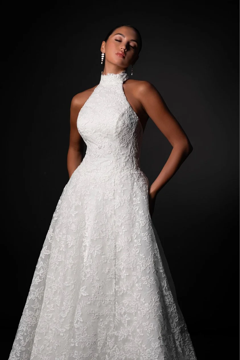 A woman wearing a white lace wedding gown with a high neckline and sleeveless design, posing against a dark background.