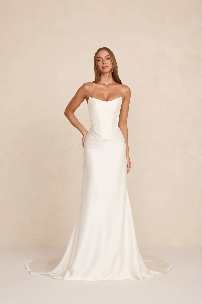 A woman in a strapless white wedding gown standing against a neutral background.