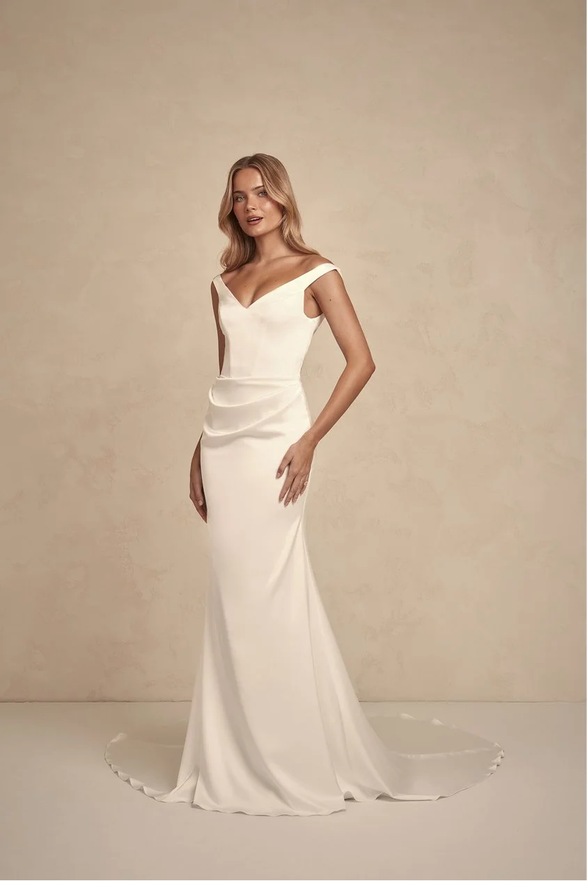 A woman wearing an elegant, long, satin wedding dress with a fitted bodice and cap sleeves, standing against a plain beige background.
