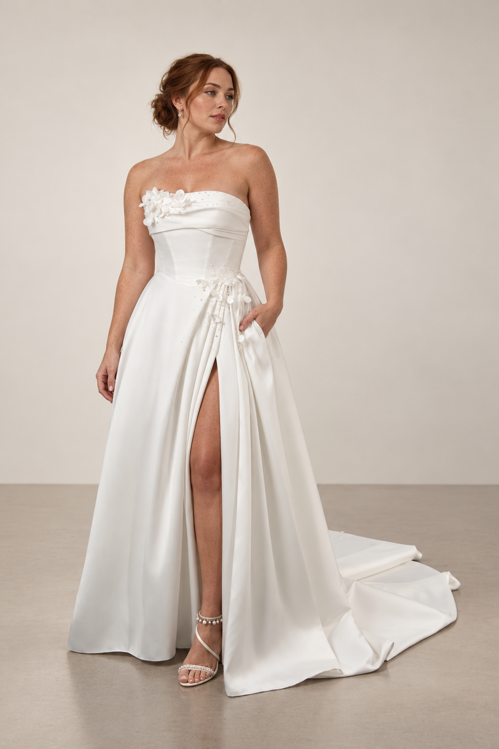 Woman in a white wedding dress with floral embellishments, standing against a plain wall, holding the side of her dress to reveal a high slit.