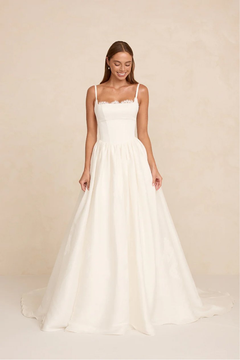 Woman in a white wedding dress with spaghetti straps, lace detail at top, smiling, standing against a beige background.