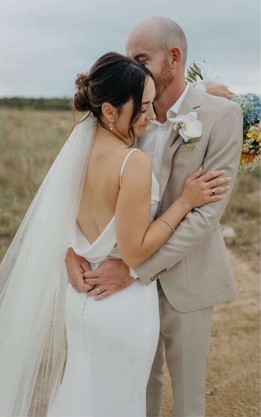 A bride and groom hugging outdoors, with the groom gently kissing the bride's forehead. The bride is wearing a white wedding dress with spaghetti straps and a veil, and the groom is in a beige suit with a white shirt and a flower boutonniere.