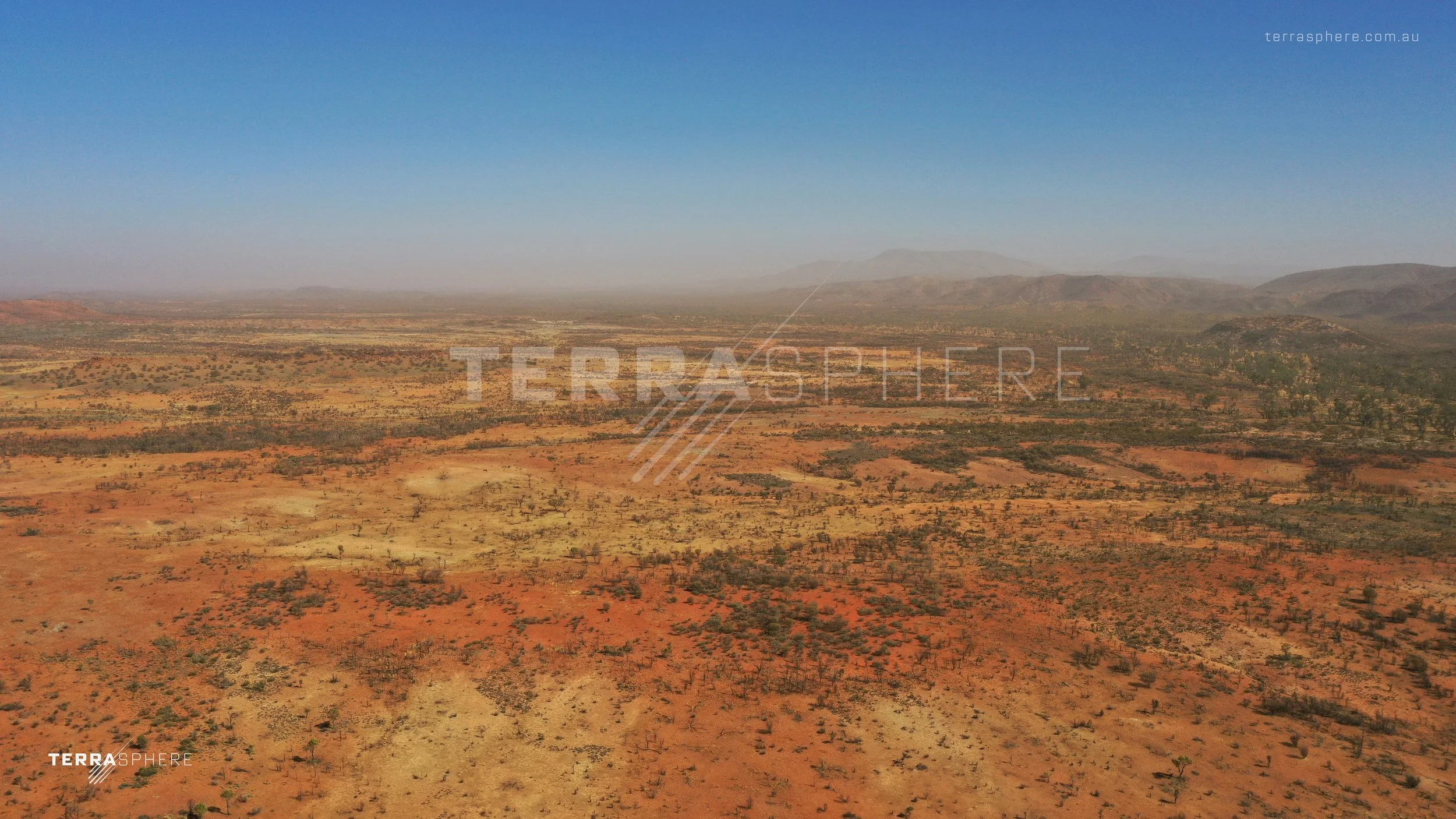 A vast arid desert landscape with sparse vegetation and distant hills under a blue sky.