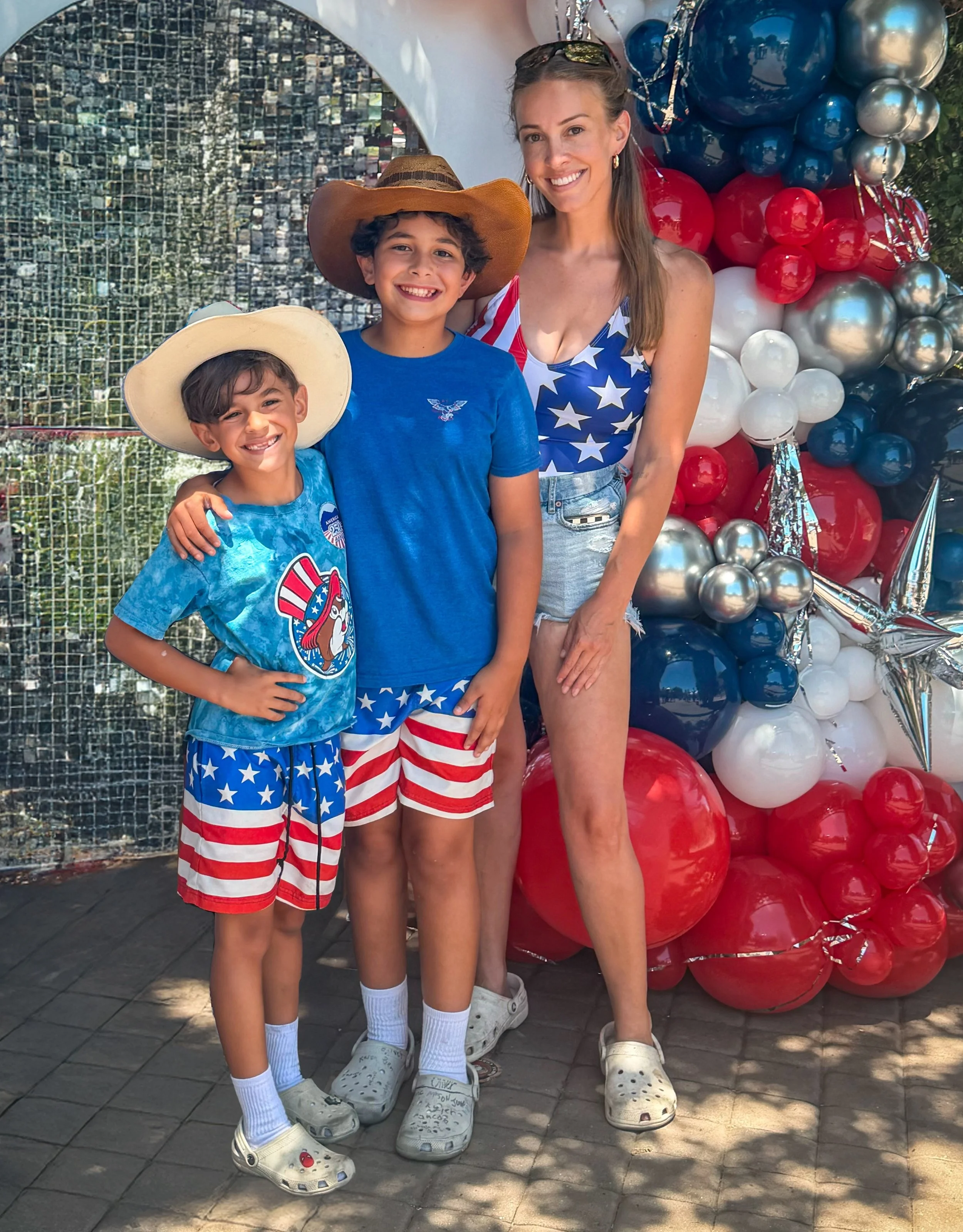 Three smiling people, two children and one woman, standing in front of red, white, blue, and silver balloons at a festive outdoor event. The children are wearing patriotic-themed outfits and matching shoes, and the woman is dressed in an American flag-themed top and denim shorts.