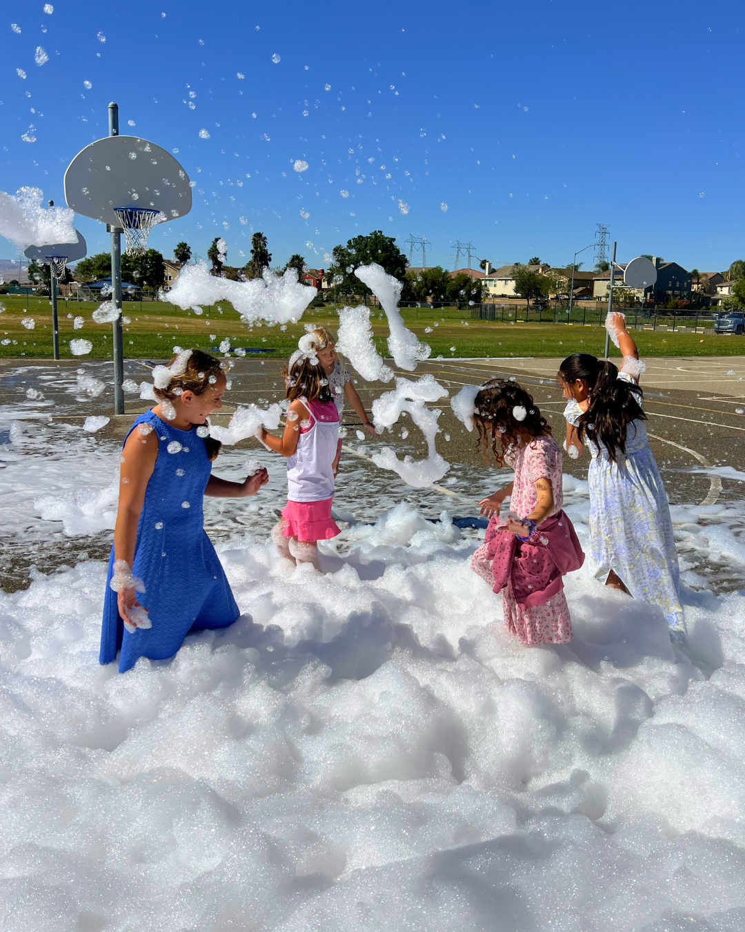 Children playing in foam on an outdoor basketball court on a sunny day.