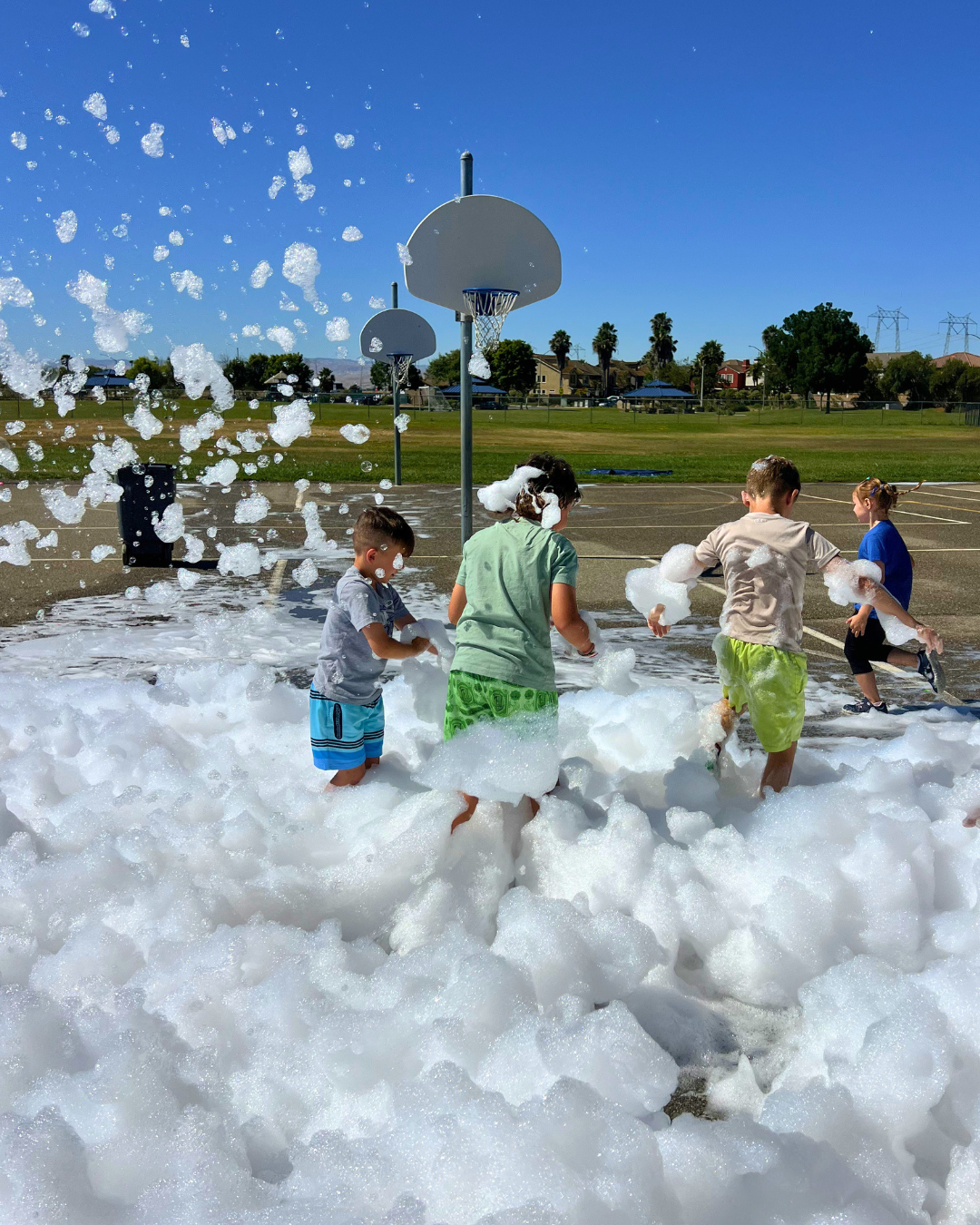 Children playing in foam on a basketball court, with a blue sky and residential houses in the background.