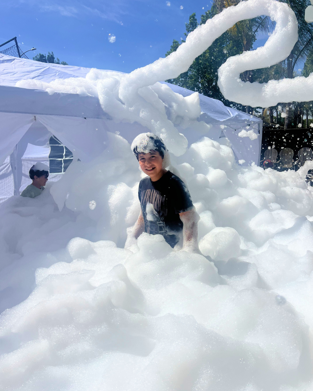 A smiling young boy standing in a backyard filled with white foam and soap bubbles, with foam foam forming a large swirl above him. In the background, there is a white canopy tent and another child inside it, and trees and a fence are visible under a clear blue sky.