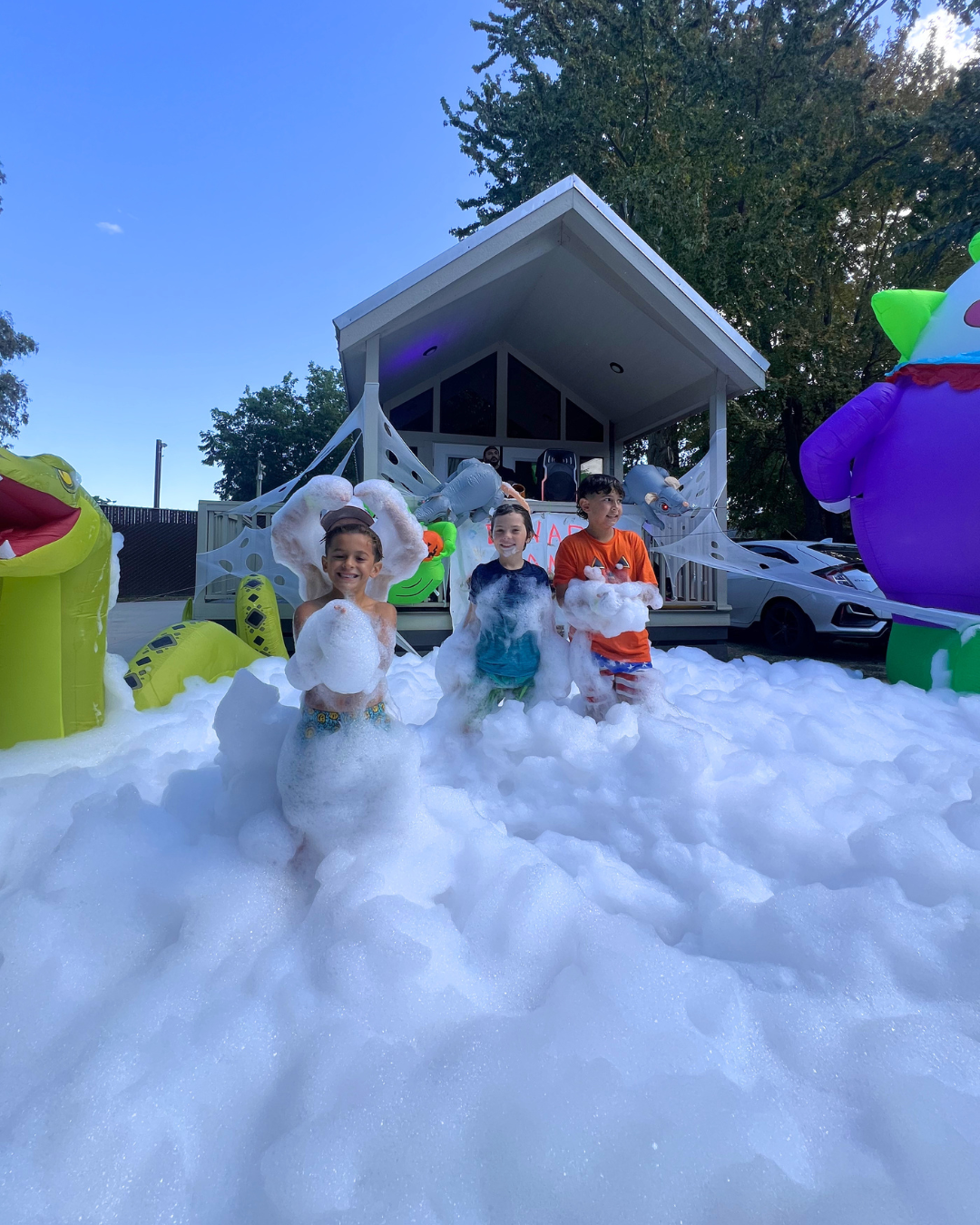 Children playing in foam bubbles in front of a house decorated with inflatable Halloween characters, including a green monster on the right and a yellow creature on the left, during a Halloween party.