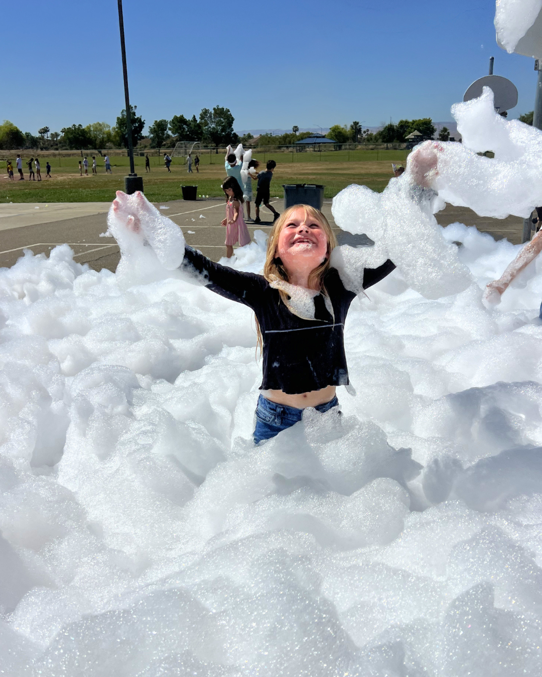 Child smiling and playing in a foam pit at an outdoor event on a sunny day, with other children in the background.