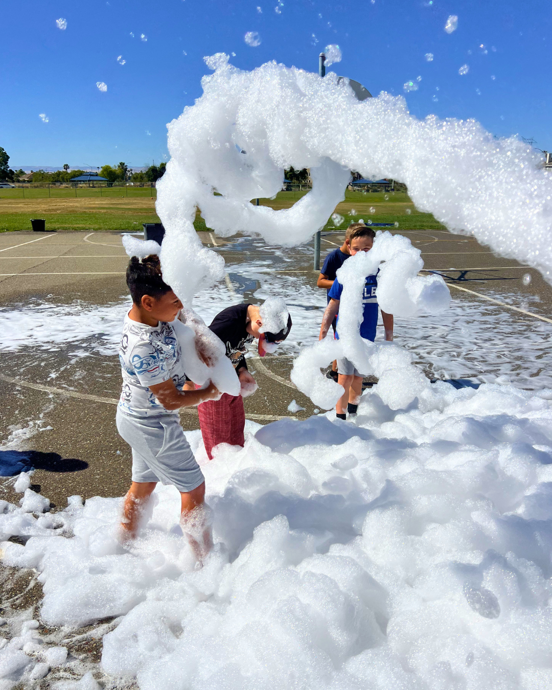 Four boys playing in a large amount of soap foam on an outdoor basketball court during a sunny day.