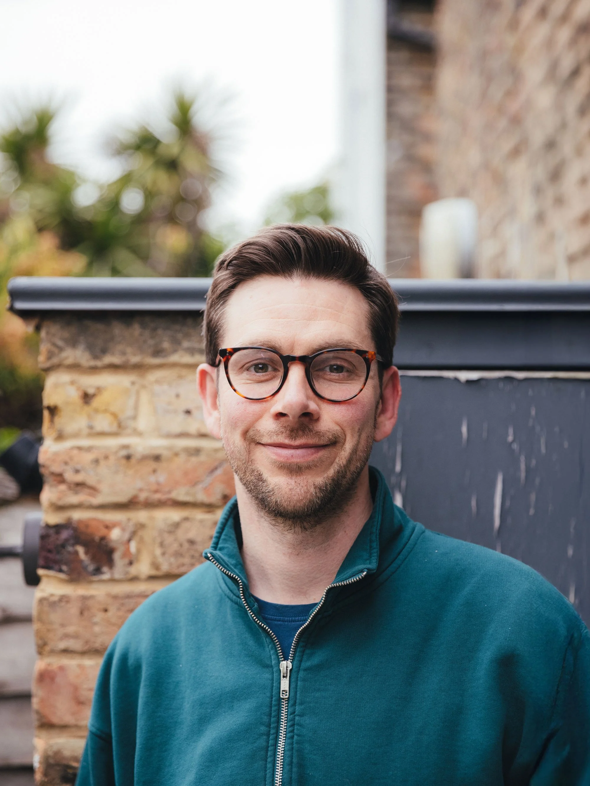 A young man with short brown hair, wearing glasses and a teal zip-up jacket, standing outdoors in front of a brick wall and a blurred background of trees.