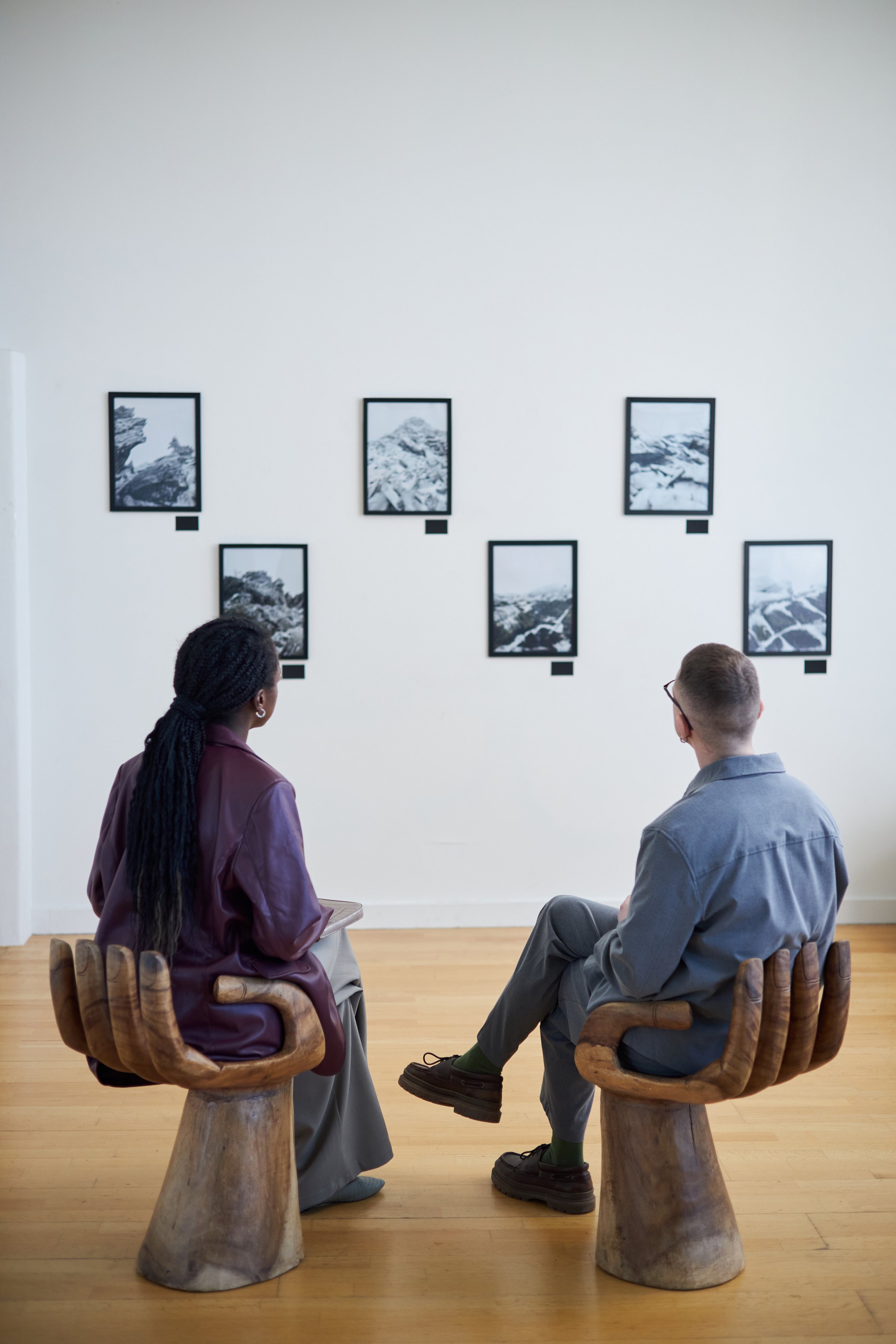 Two people sitting on wooden chairs in an art gallery, facing a wall with six framed black-and-white photographs of mountain landscapes.