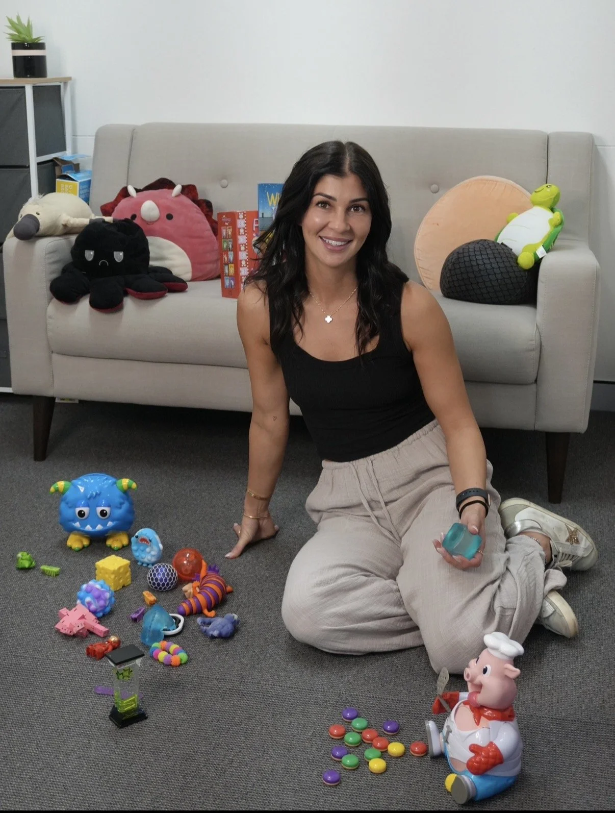 Woman sitting on carpeted floor surrounded by colorful toys, smiling at camera, with a light gray sofa and plush toys in the background.